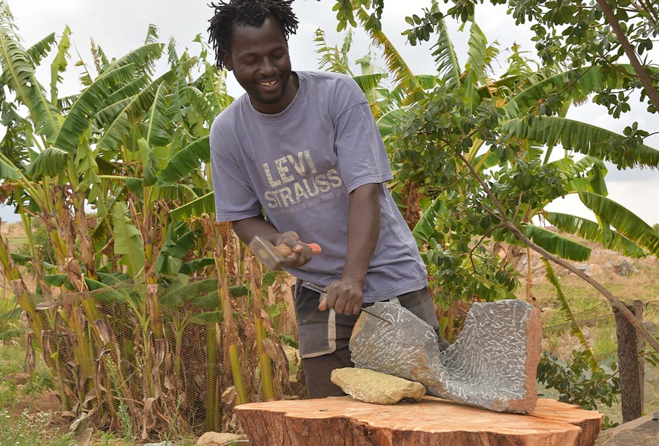 Ein afrikanischer Mann bearbeitet einen Stein auf einem Holzstamm im Freien mit Hammer und Meißel